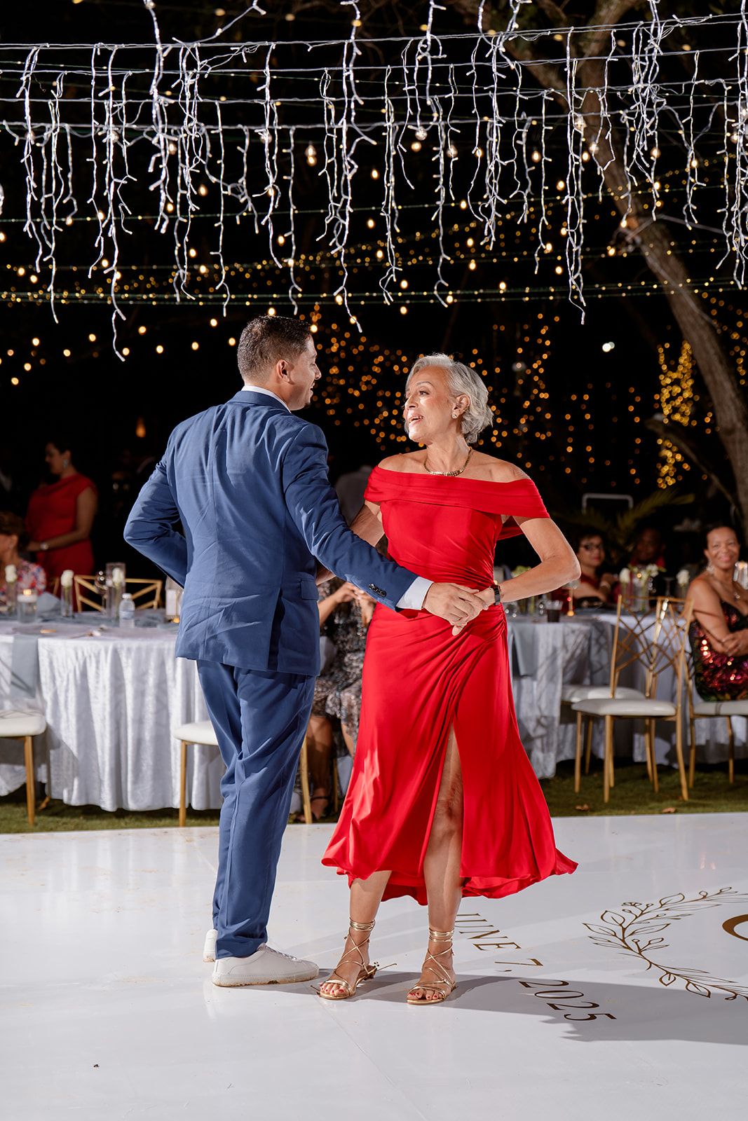 A couple dancing at an elegant outdoor reception under hanging string lights with white tables and decor in the background.