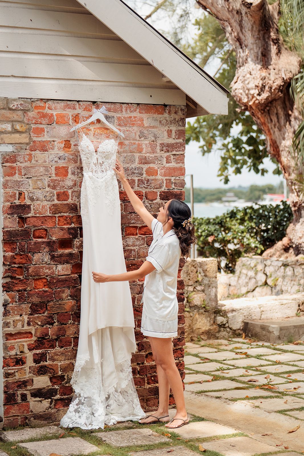 Wedding dress hangs from rustic brick wall archway in outdoor garden setting with stone pathway and greenery.