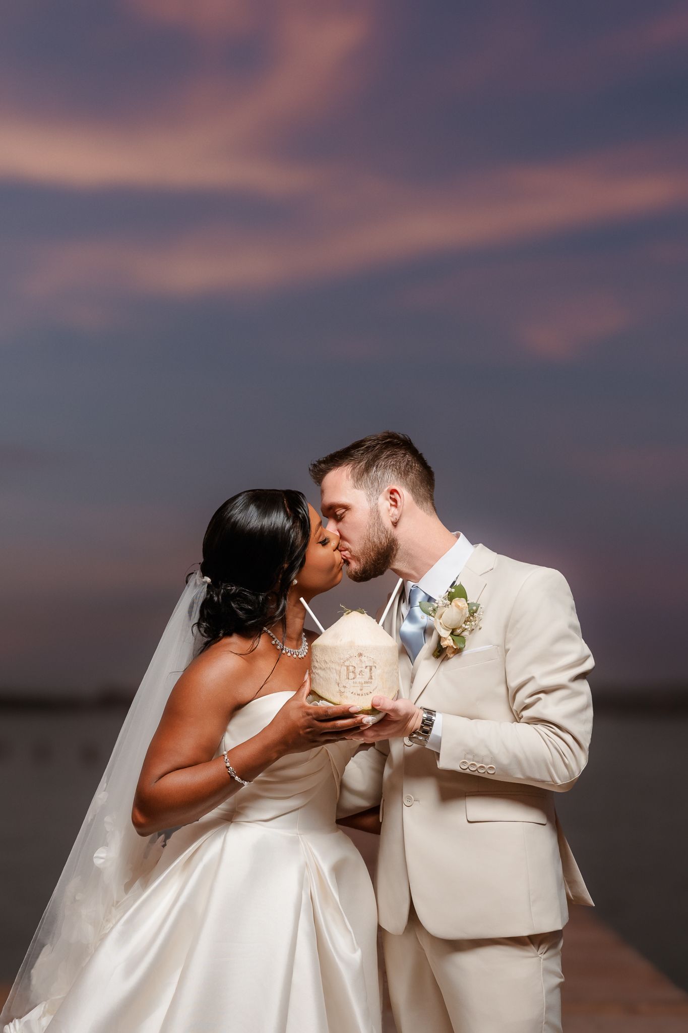 Bride and groom share a kiss at sunset while holding a coconut drink, dressed in elegant white wedding attire.