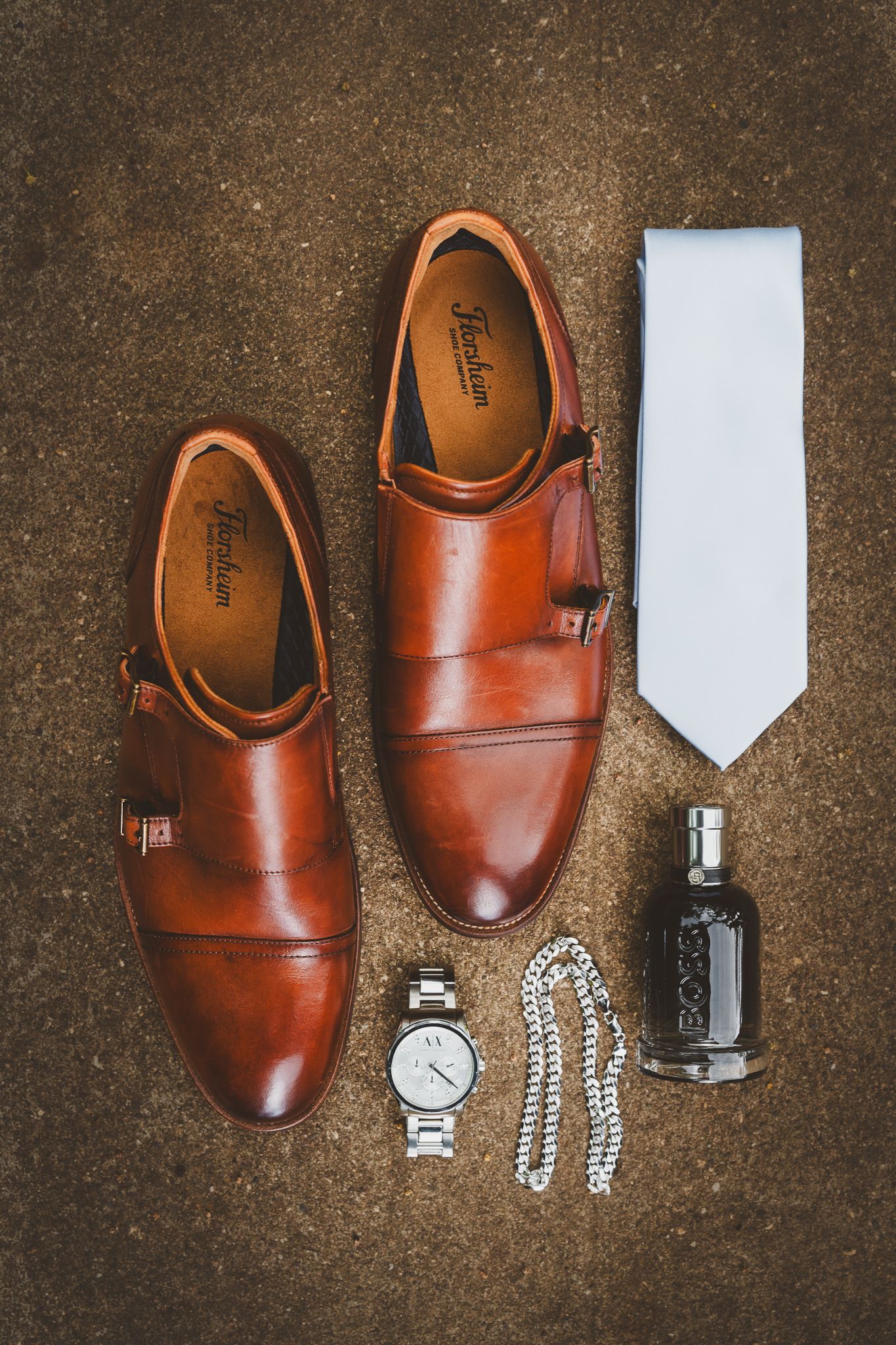 Brown monk strap dress shoes displayed with white tie, silver watch, chain necklace, and Hugo Boss cologne on stone surface.