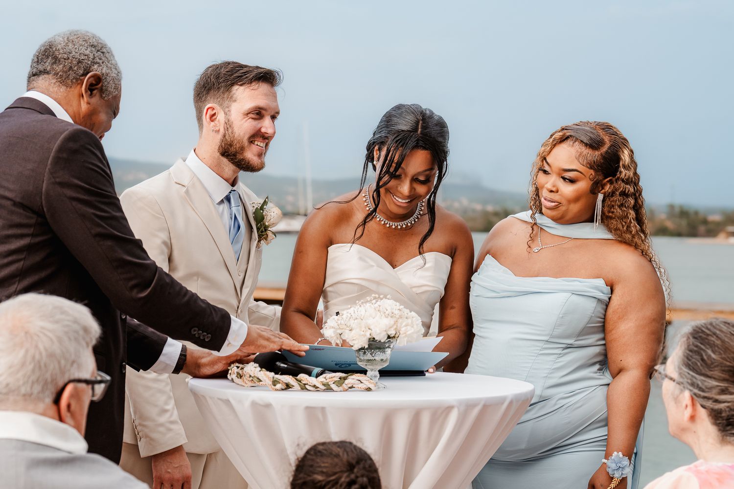 Interracial couple signs wedding documents outdoors with bridesmaid in light blue dress during waterfront ceremony.