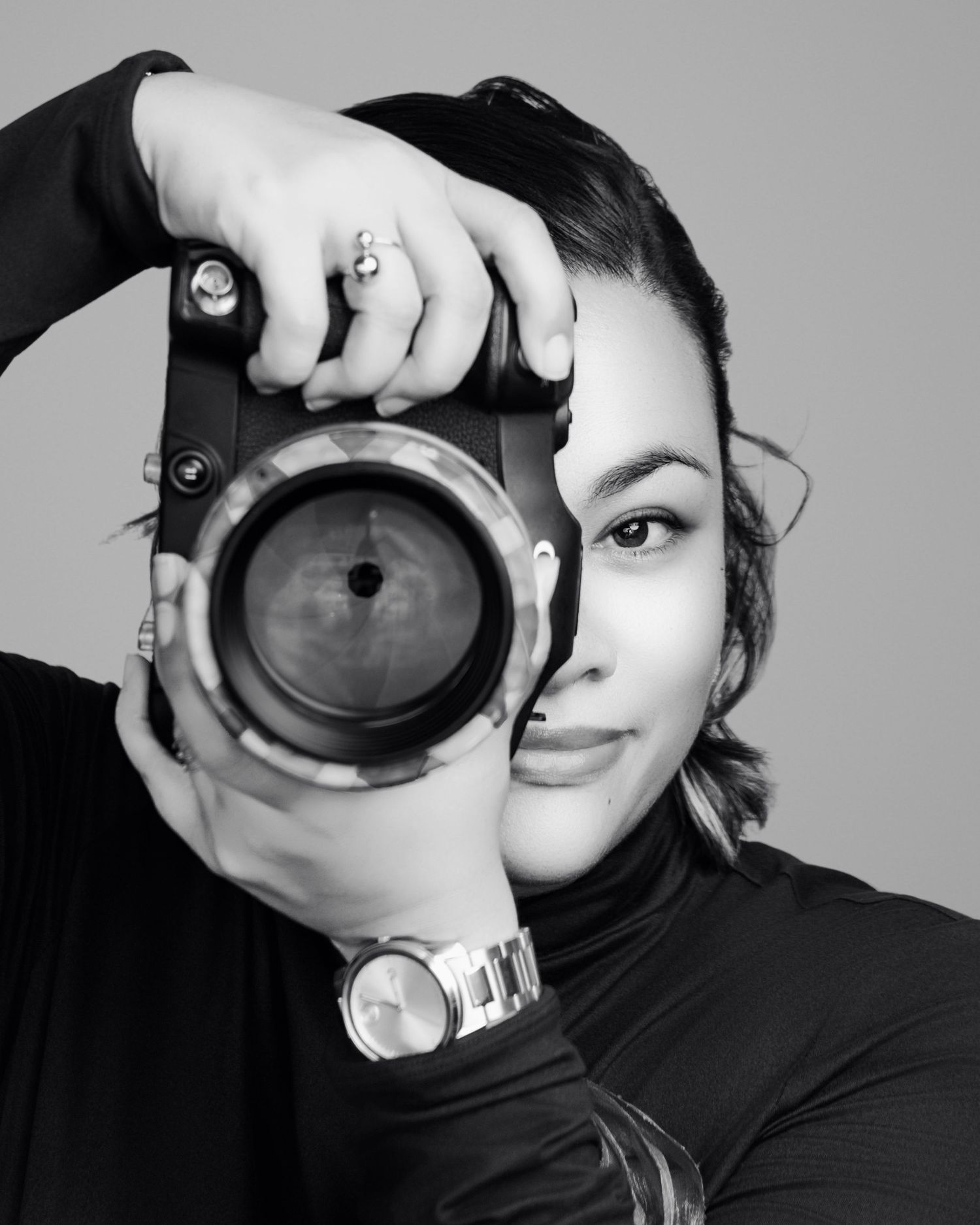 Photographer holds up a black camera lens in front of their face, creating an artistic black and white composition.