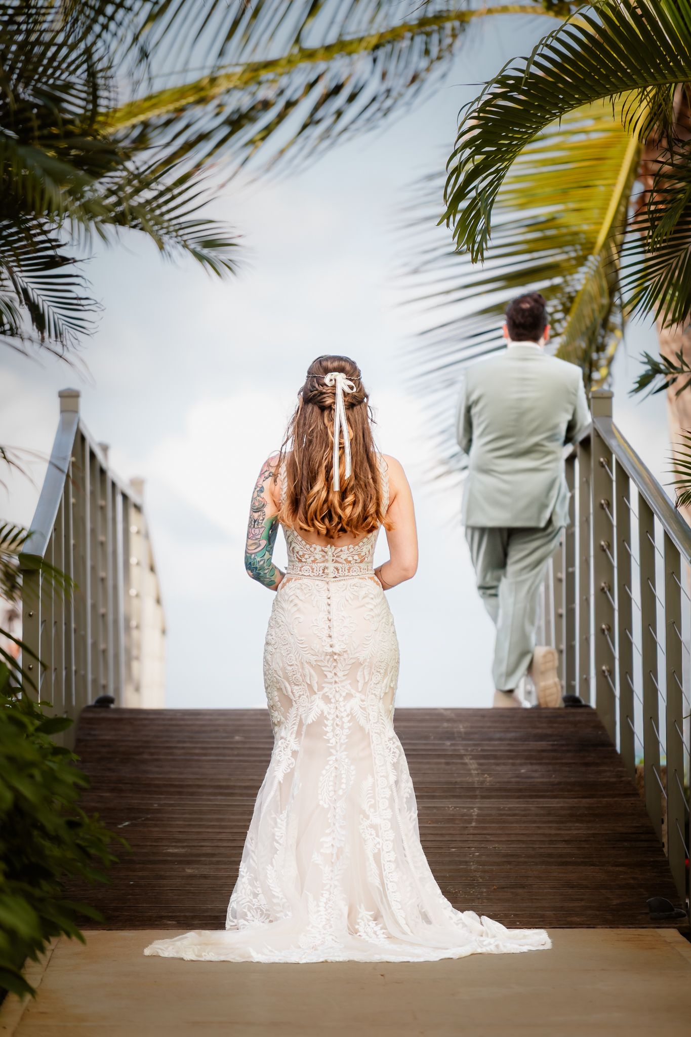 Bride in lace mermaid gown stands on wooden boardwalk steps as groom walks ahead, framed by tropical palm trees.