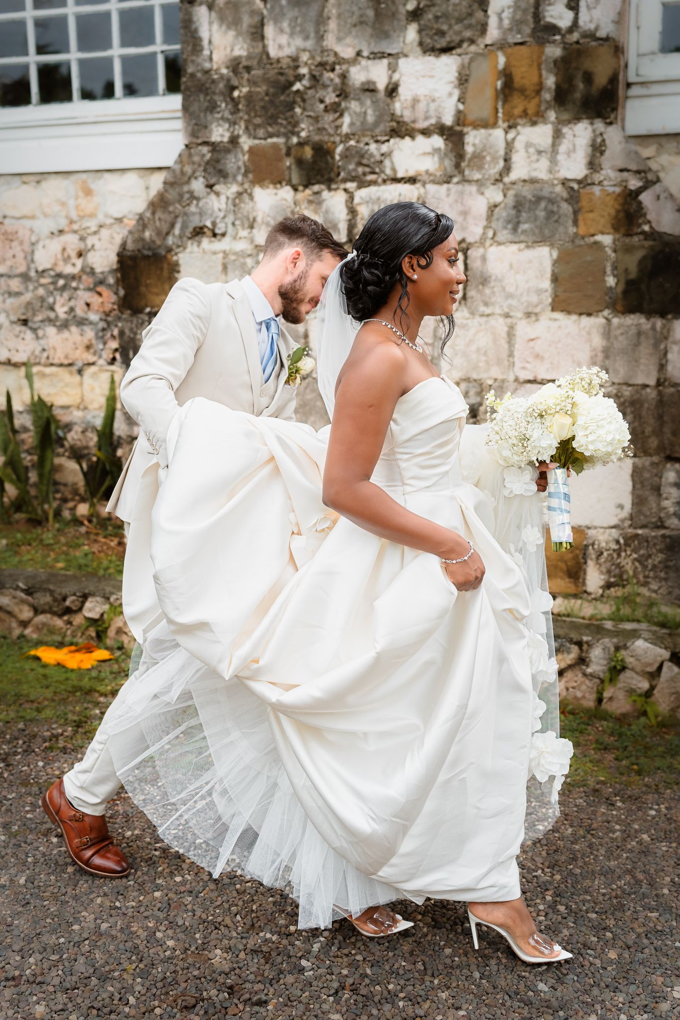 A bride in a strapless white gown holds a bouquet while her groom in a cream suit lifts her dress train as they walk.