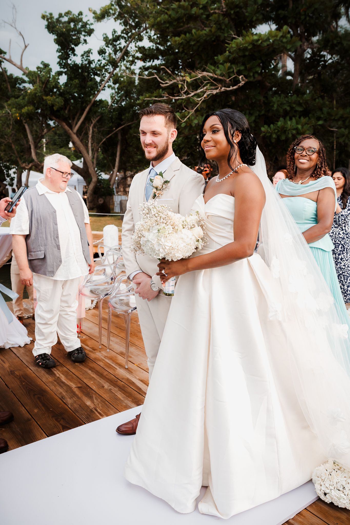 Bride in strapless white gown holding bouquet walks with groom in tan suit at outdoor wedding ceremony.