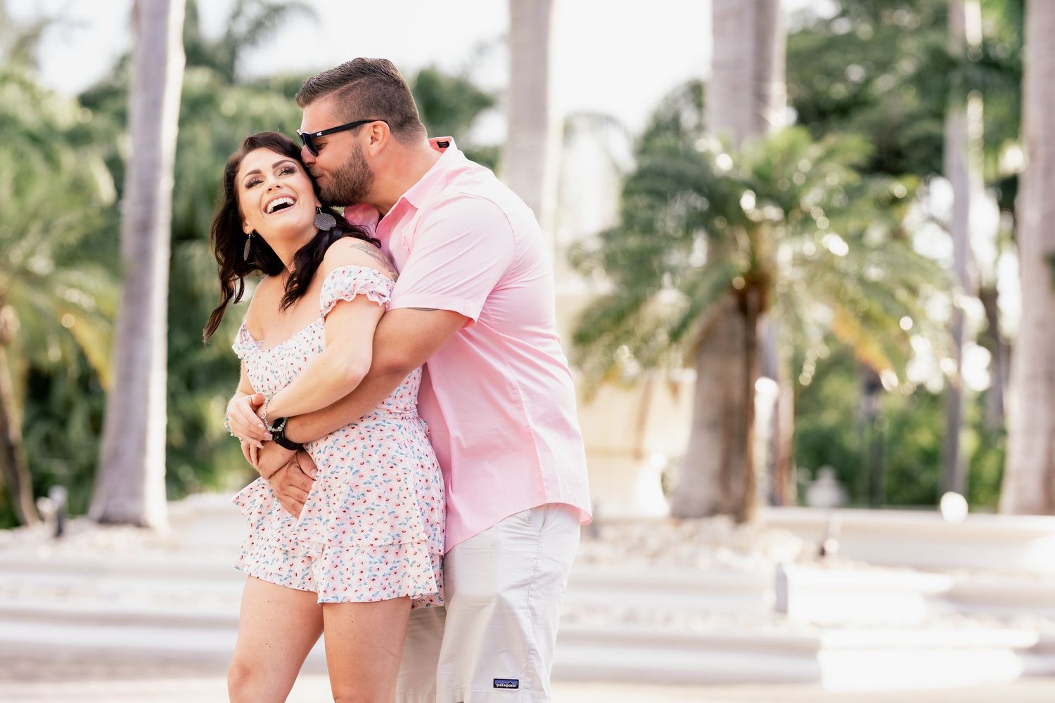 A couple embraces and laughs together in a tropical setting with palm trees in the background.