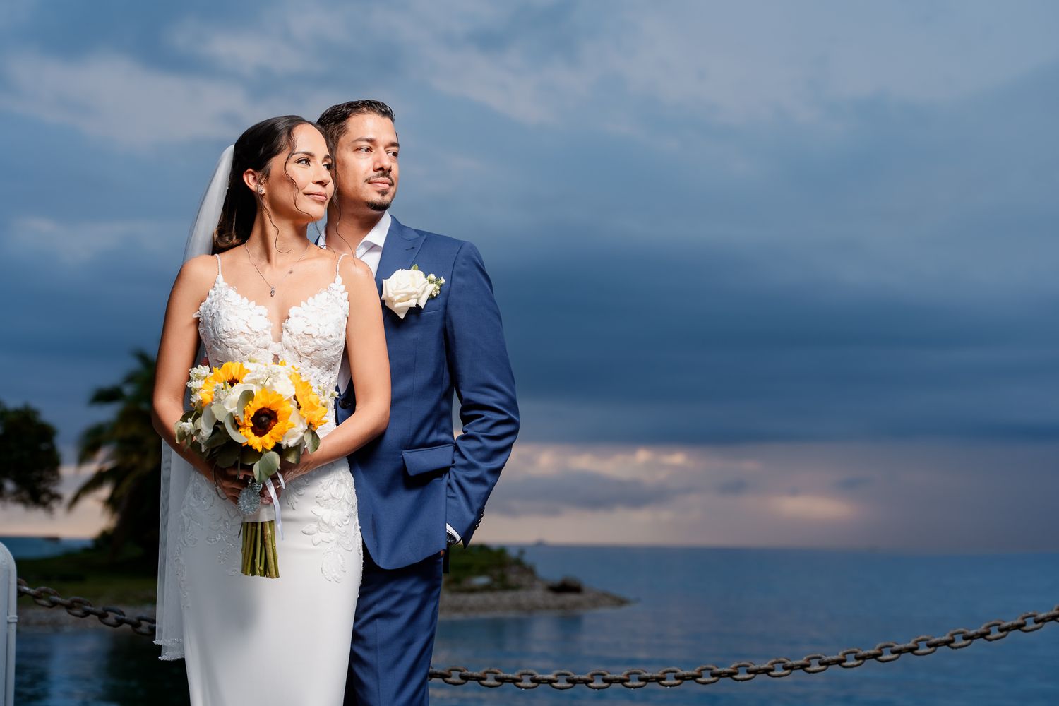 A couple poses at sunset by the ocean, with sunflowers in a bridal bouquet and a chain barrier nearby.