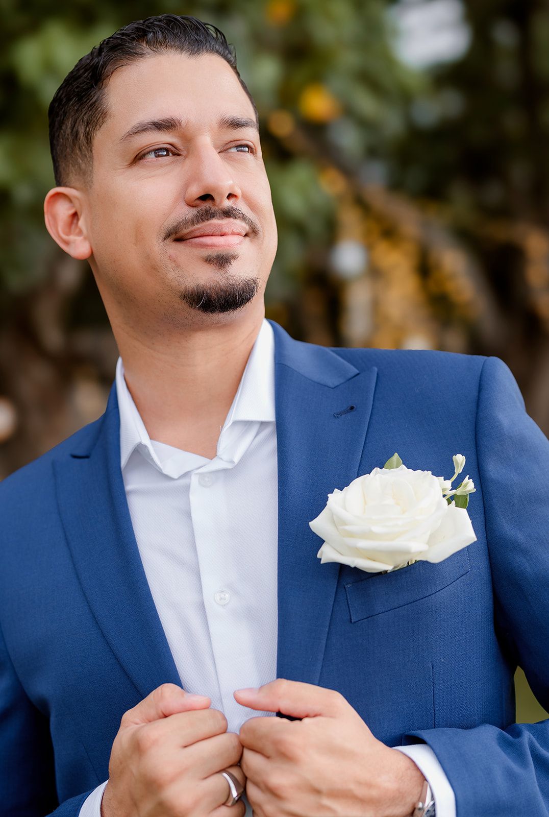 Groom in navy blue suit with white boutonniere looks upward with smile during outdoor wedding photography session.