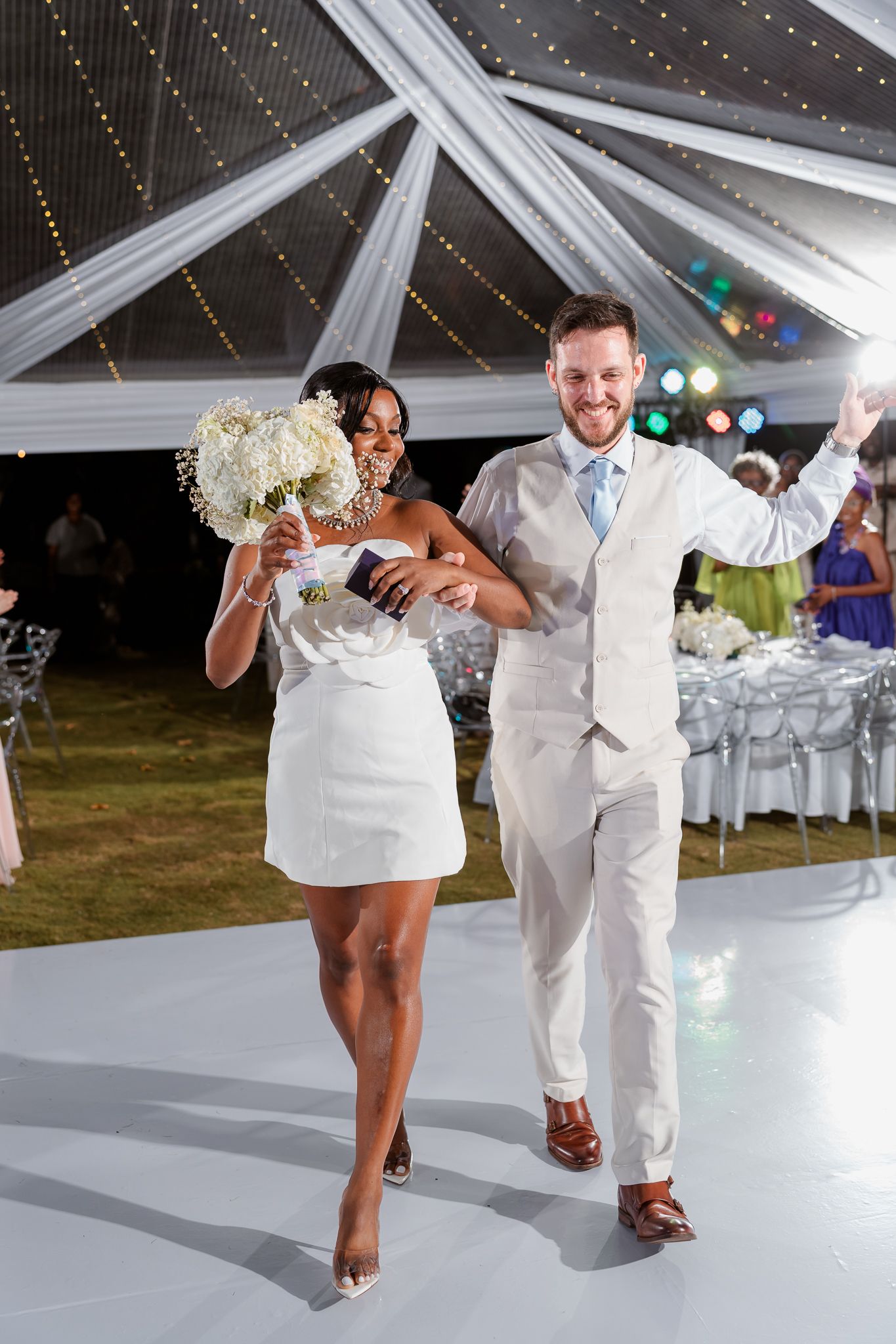 Interracial couple in wedding attire celebrating on dance floor under decorated tent, bride holding bouquet.