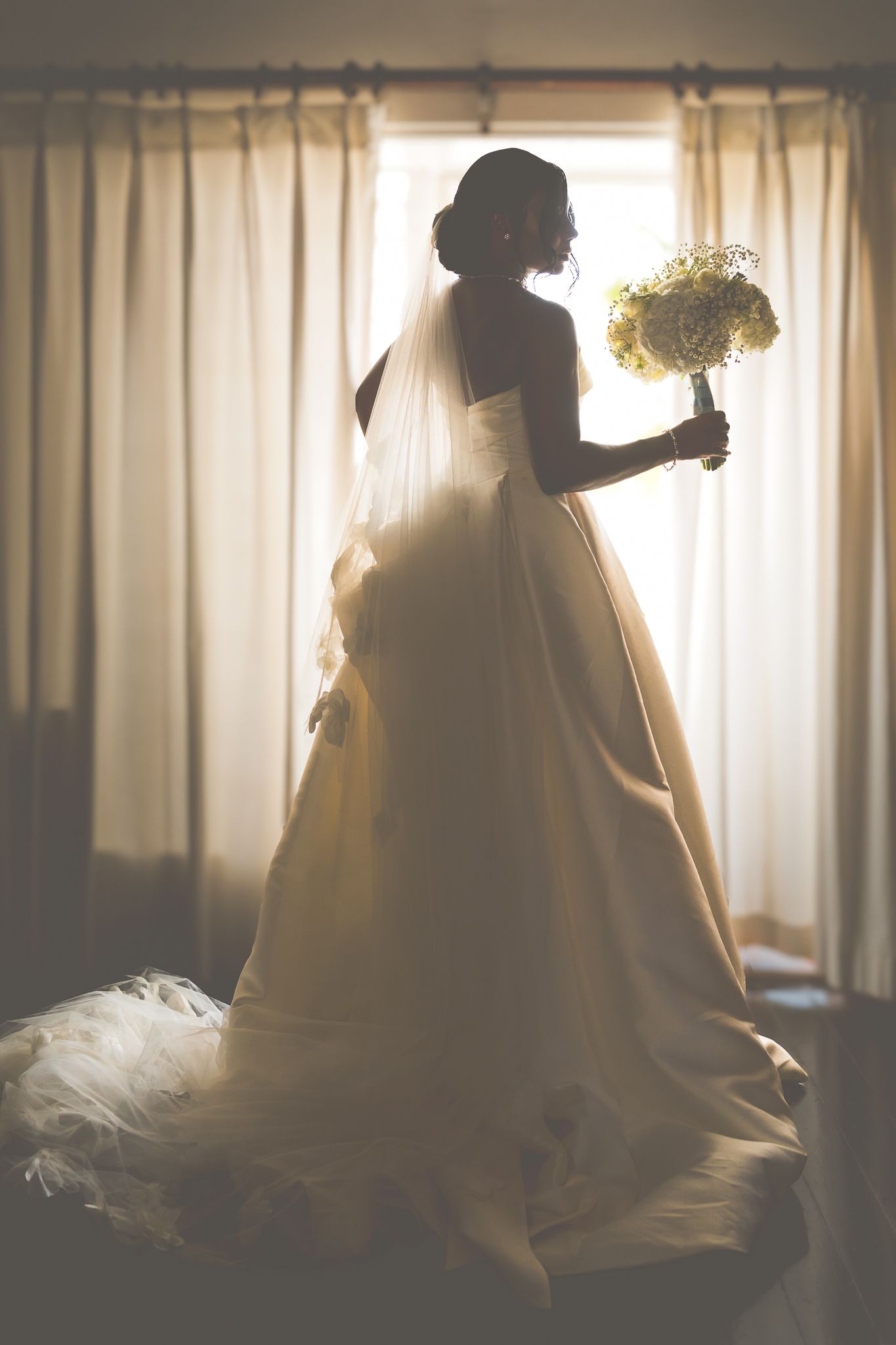 Bride in elegant white wedding gown and veil holding bouquet, silhouetted against bright window with curtains.