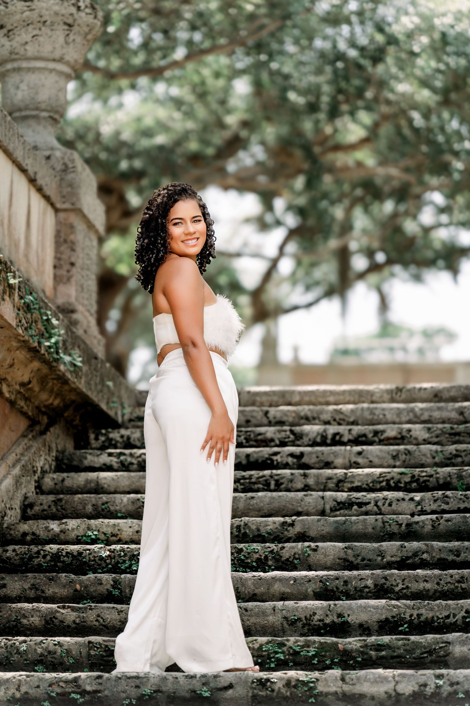 A figure in a flowing white dress poses gracefully on stone steps surrounded by lush trees and natural scenery.