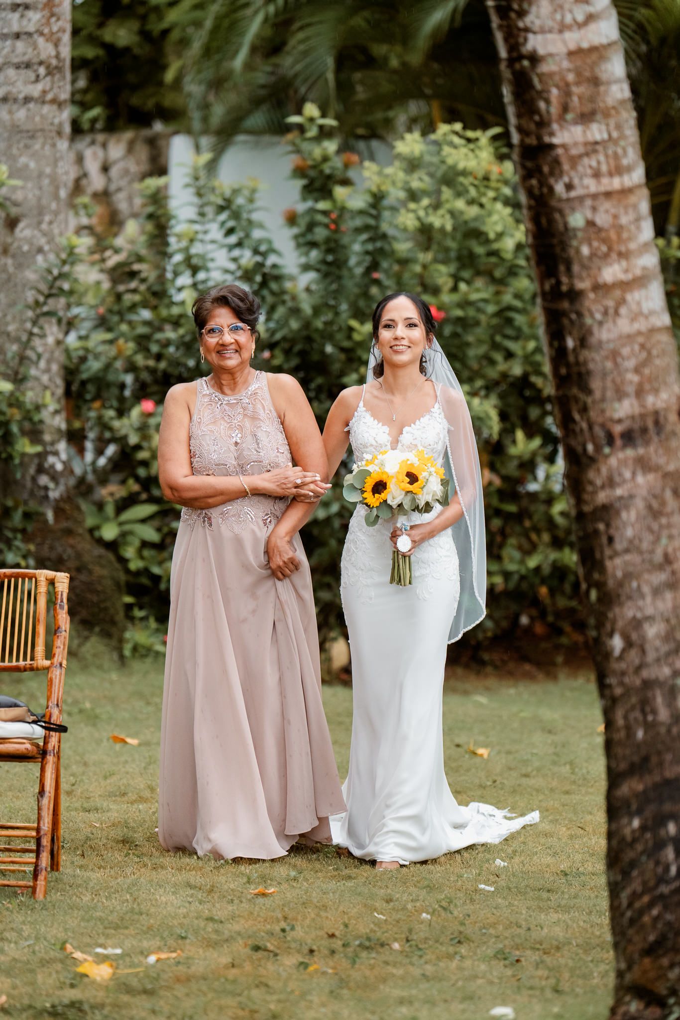 Two women in formal dresses stand on grass under palm trees, with one holding a sunflower bouquet at an outdoor wedding.