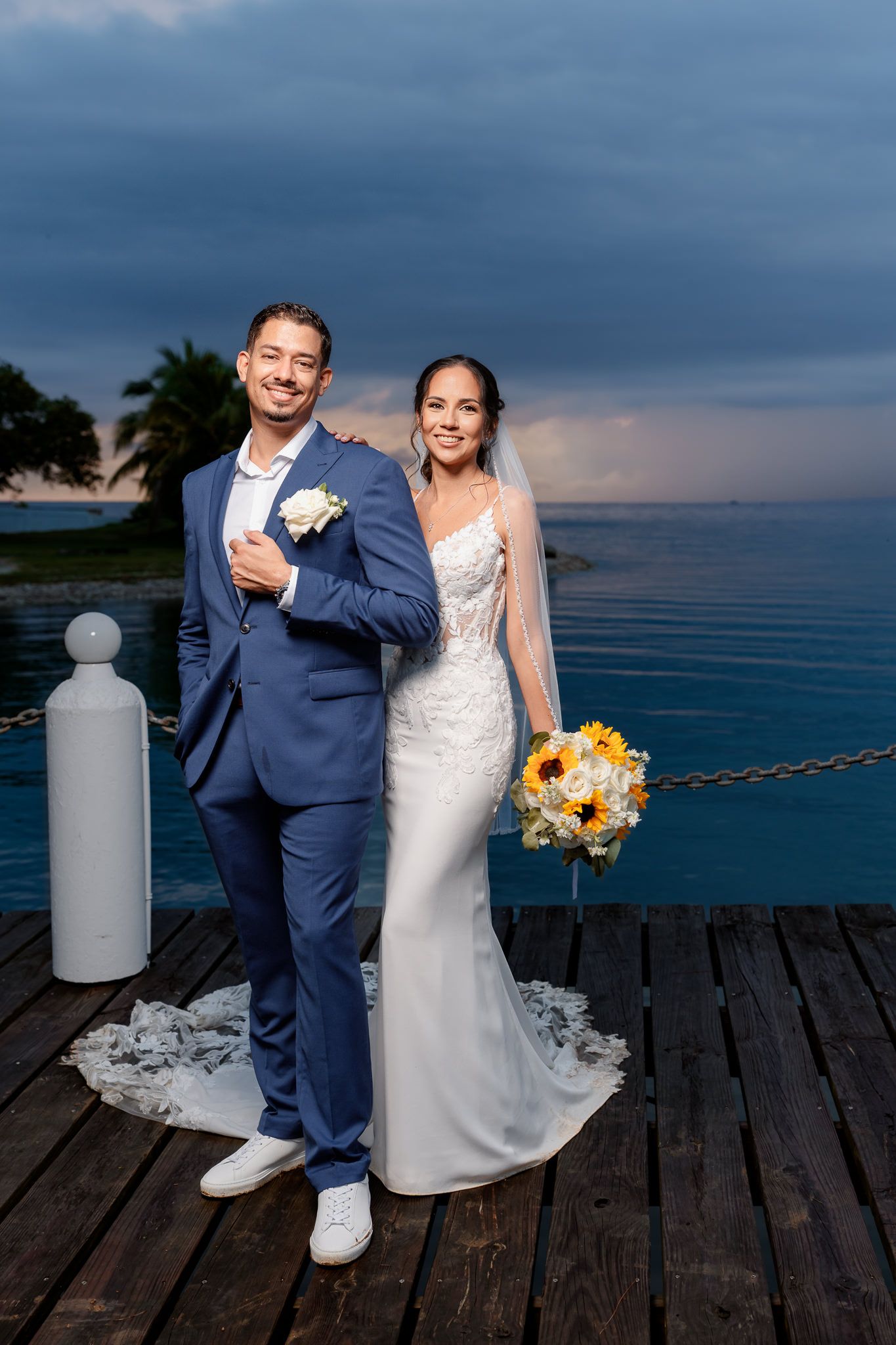 A couple poses on a wooden deck overlooking the ocean at sunset during their beach wedding celebration.