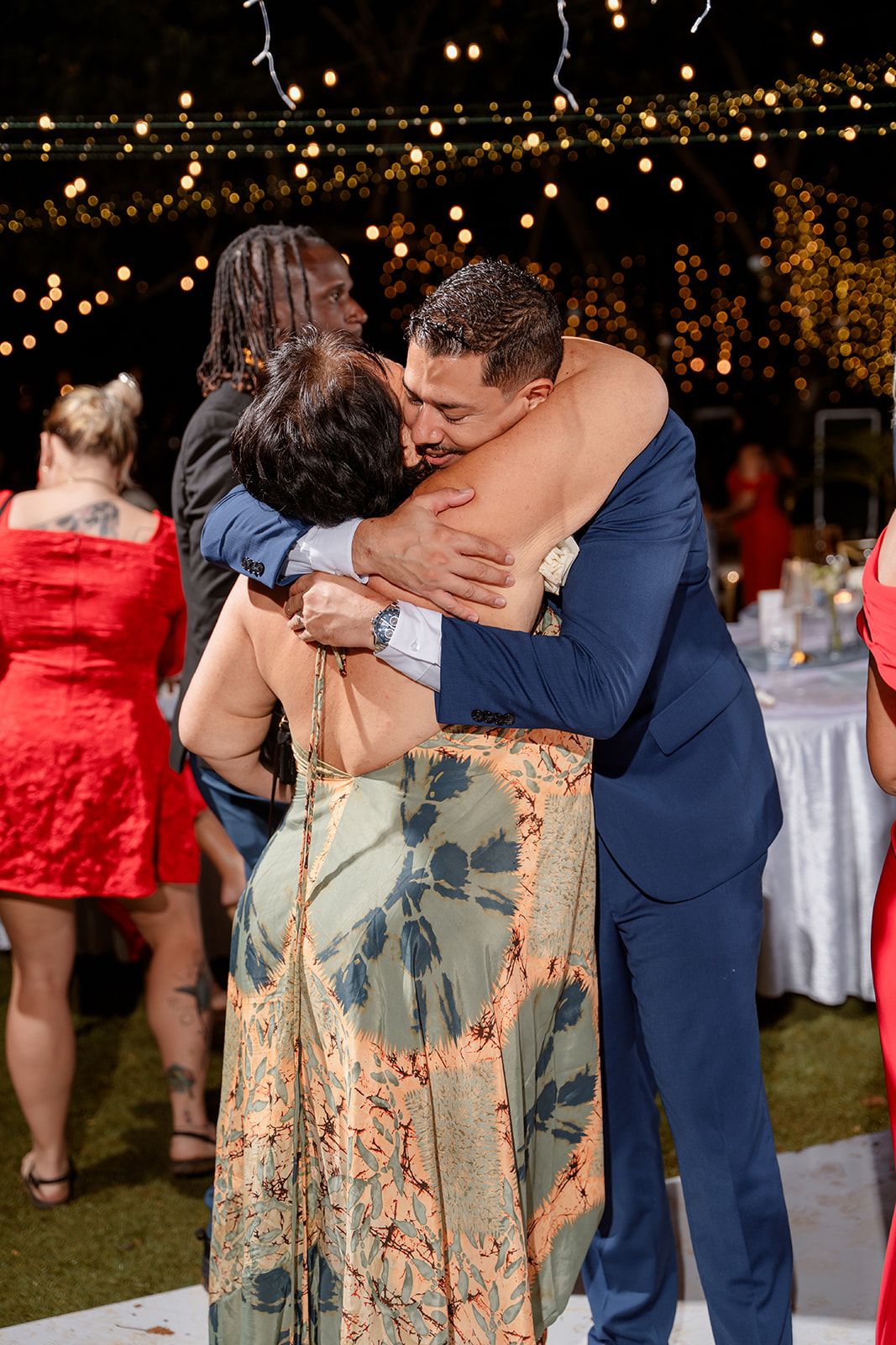 Two people embracing warmly at an outdoor evening reception with twinkle lights and decorations in the background.