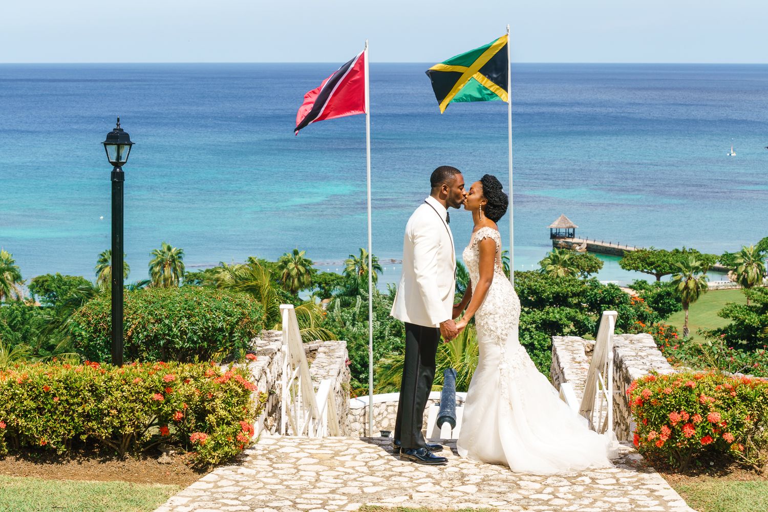 Bride and groom kissing on stone bridge with Jamaican and Trinidadian flags, overlooking turquoise Caribbean sea.
