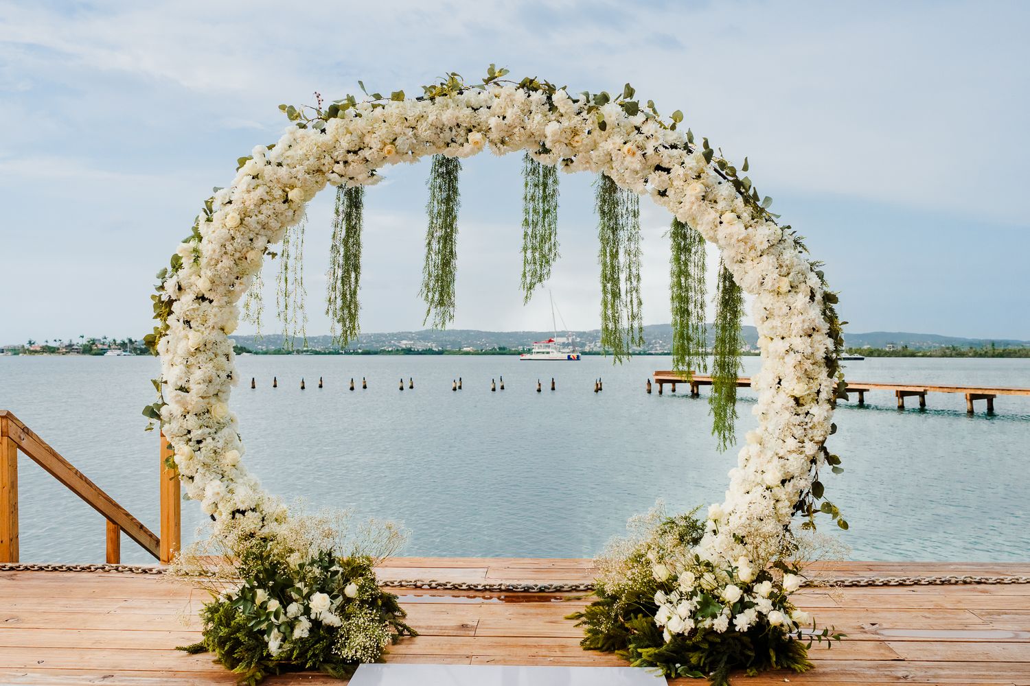 Elegant circular floral arch with white flowers and greenery on a waterfront dock for an outdoor wedding ceremony.