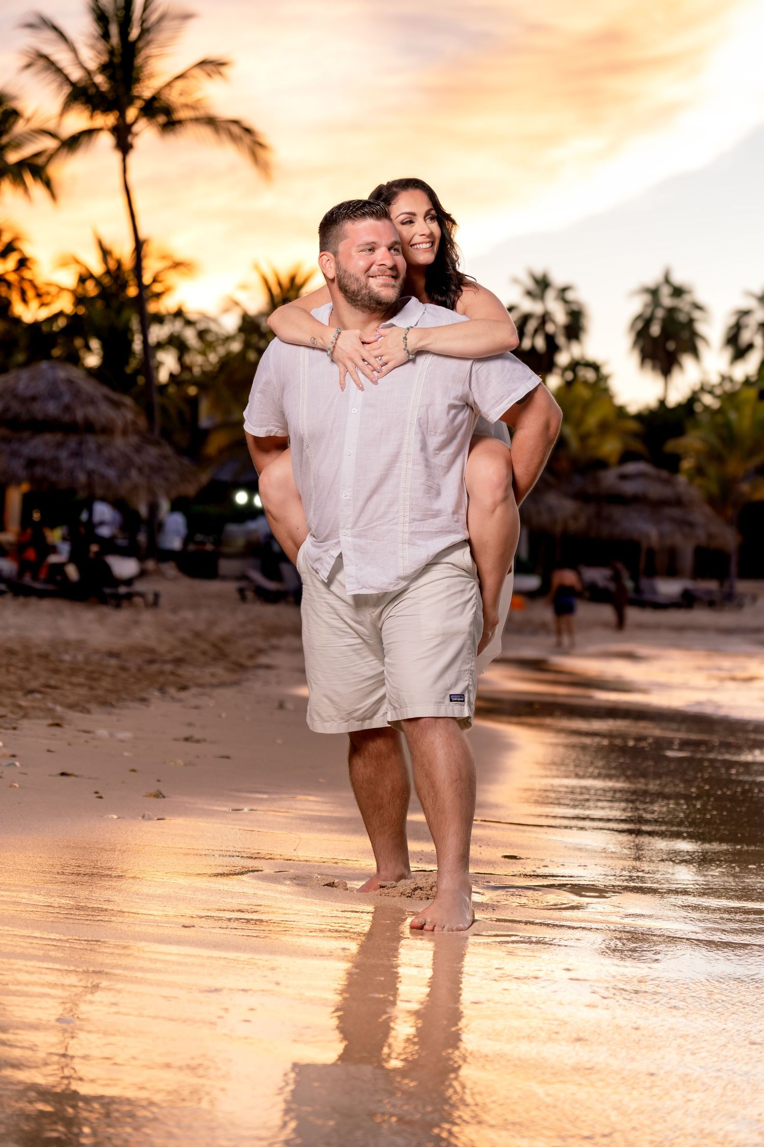 A romantic couple embraces on a tropical beach at sunset with palm trees and thatched huts in the background.
