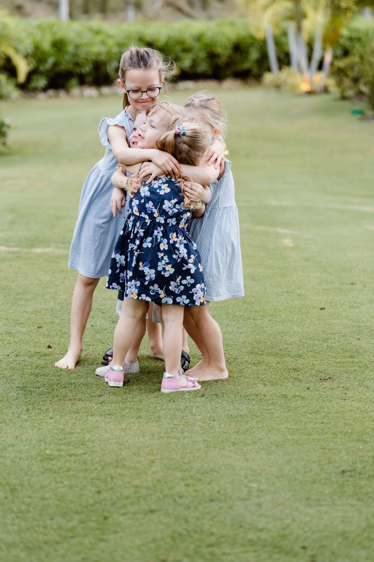 Young children in summer dresses share a joyful embrace while playing barefoot on green grass.