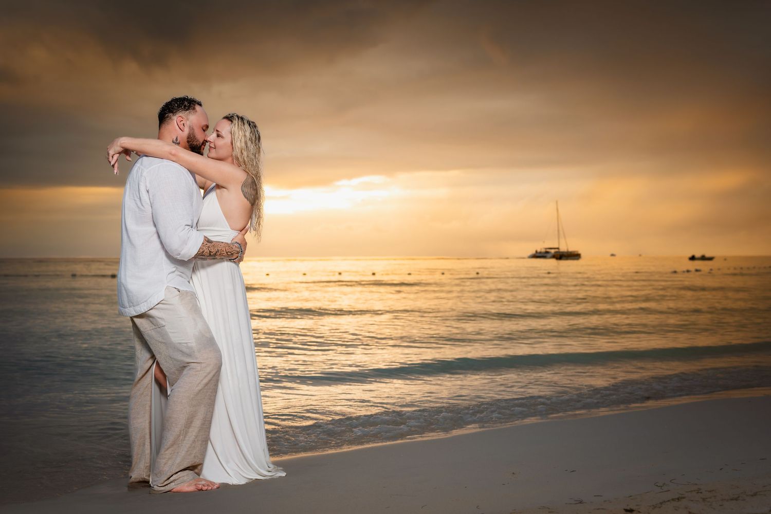 Couple sharing a romantic kiss on a sandy beach at golden sunset with calm ocean and sailboat in background.