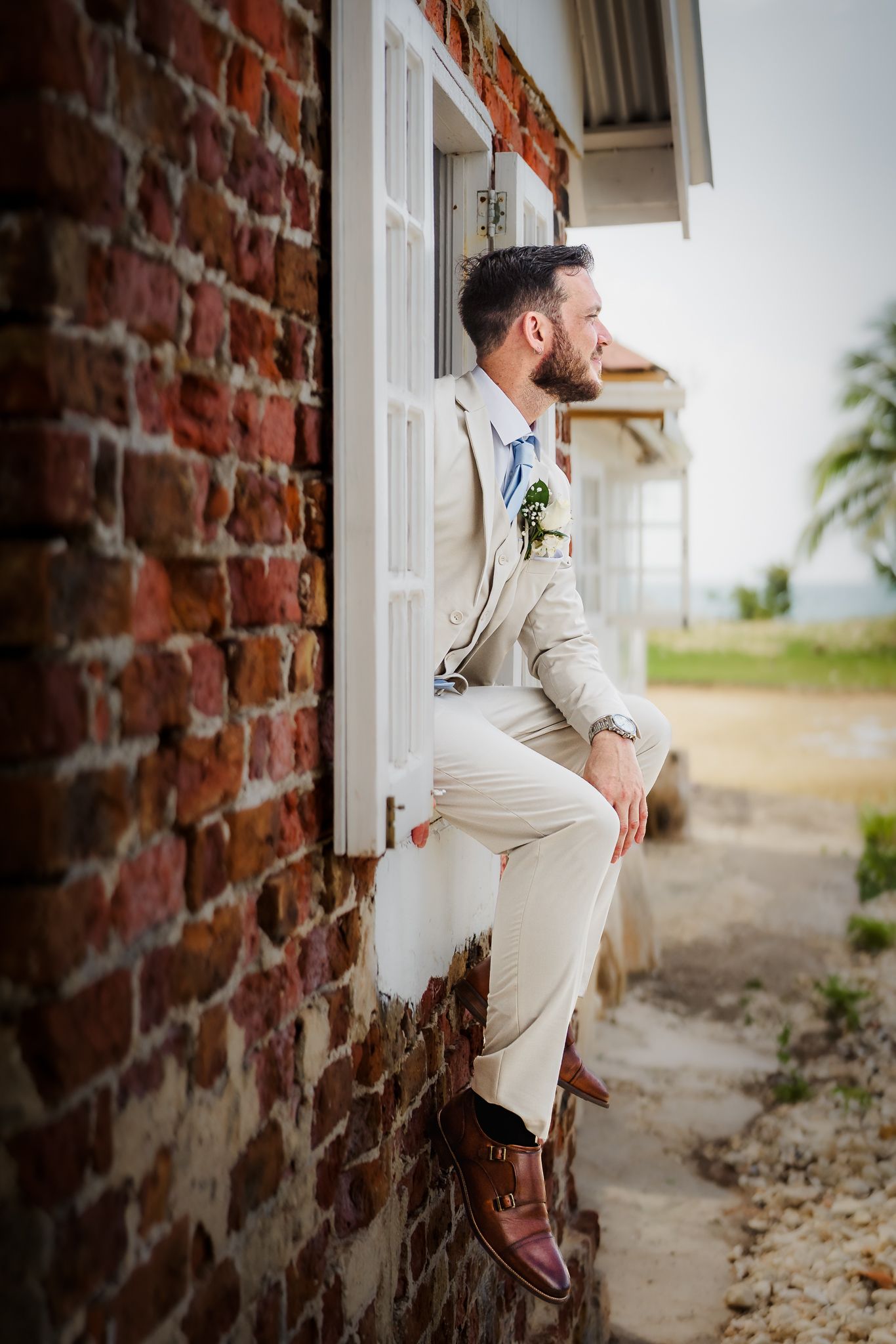 Bearded groom in beige suit sitting on windowsill of rustic brick building, wearing brown monk strap shoes and boutonniere.
