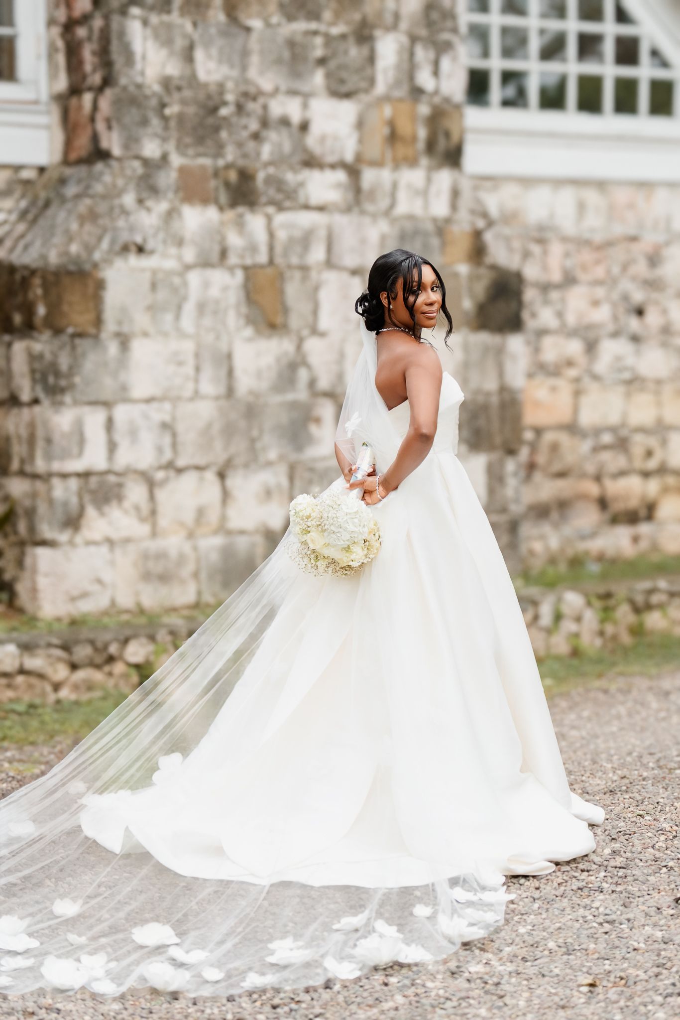 Bride in elegant white ballgown with floral veil train holds bouquet before stone building.