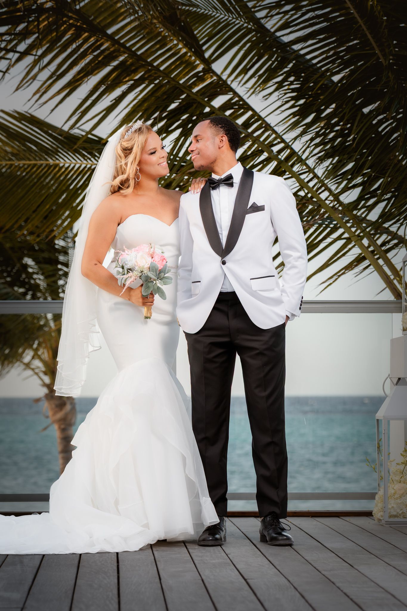 Bride and groom posing on a tropical waterfront deck with palm trees, bride holding a bouquet in a white mermaid gown.