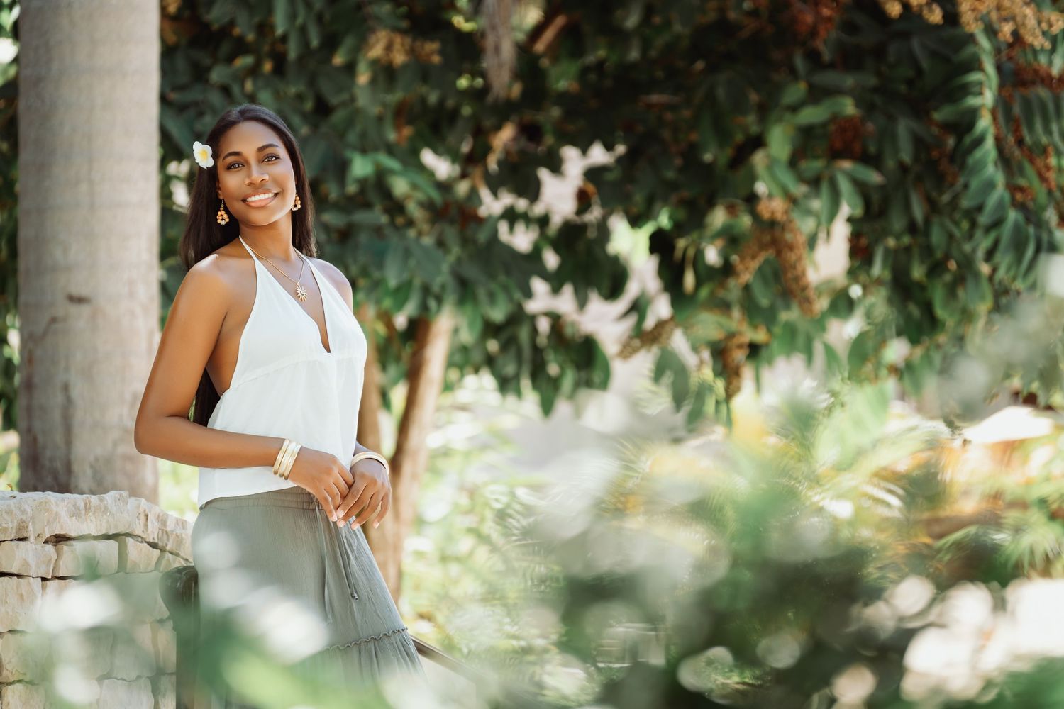 Woman in white dress poses gracefully against stone pillars in a lush garden setting.