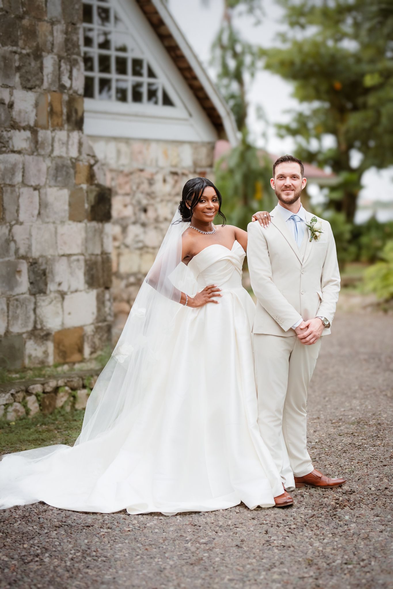 Bride in white strapless gown with veil and groom in beige suit pose together outside a stone building.