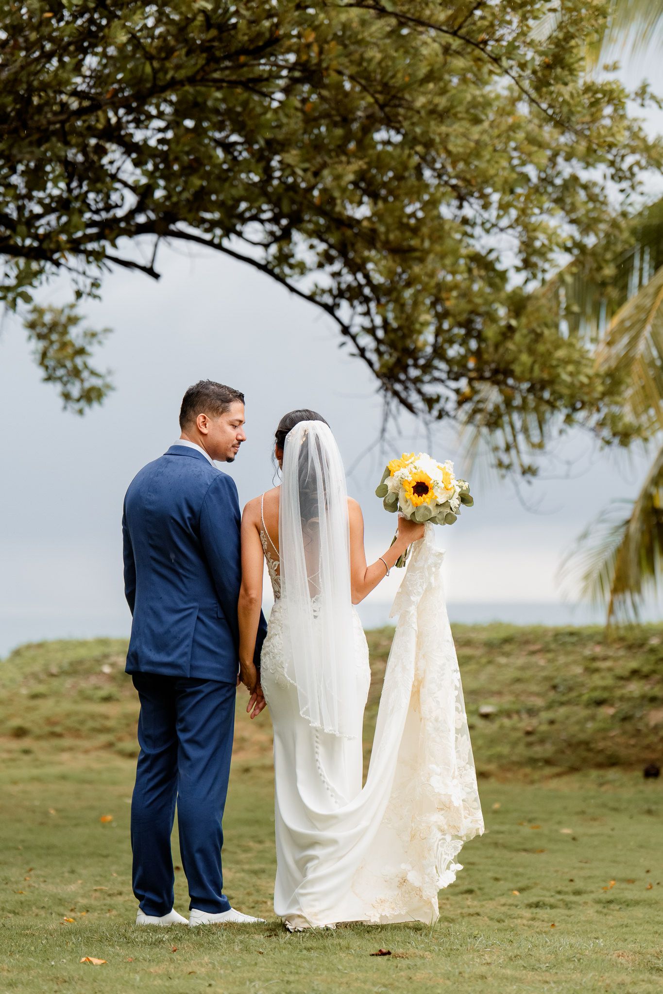 A couple in wedding attire stands outdoors with the bride holding a yellow bouquet under a tree canopy.
