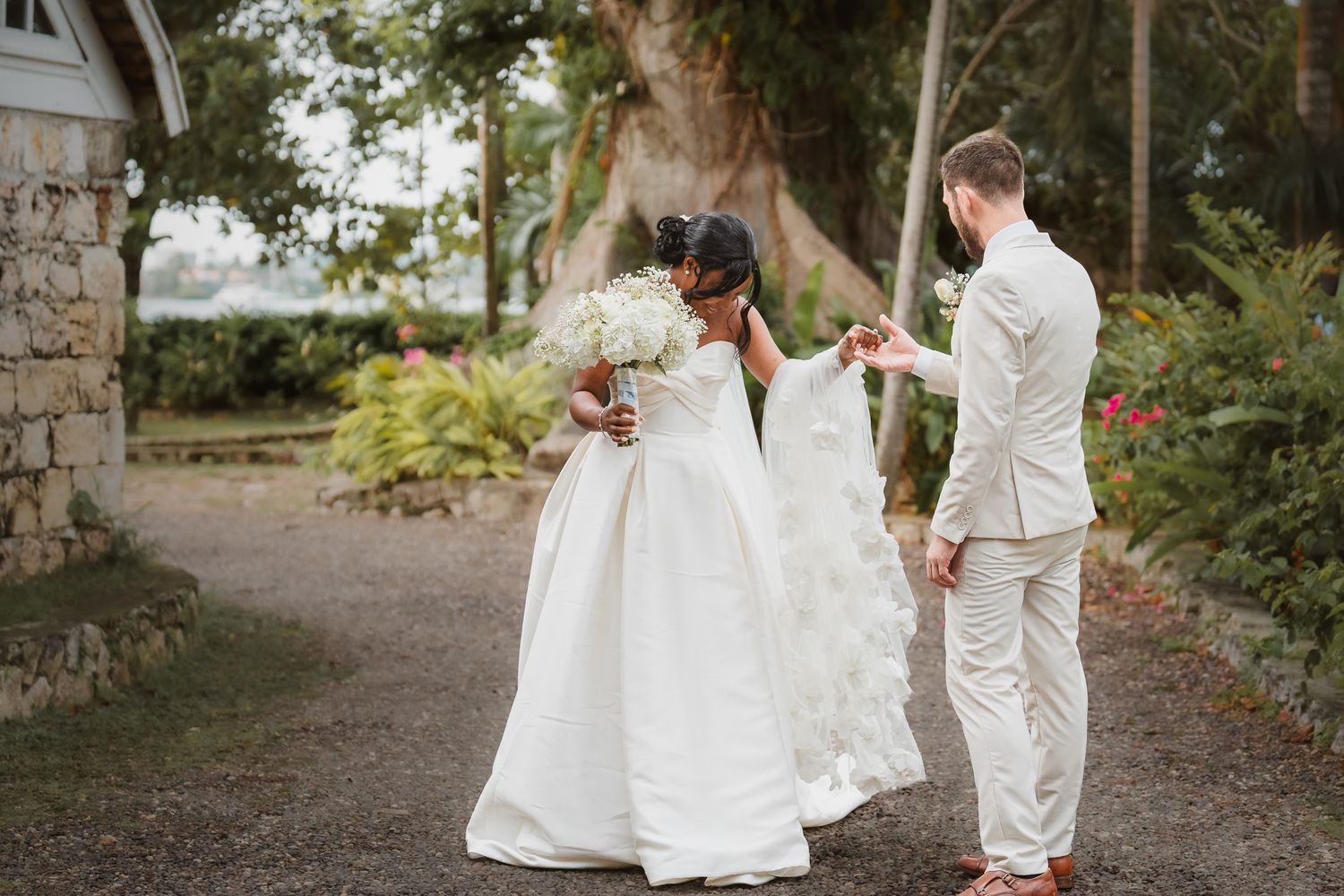 Bride in white gown holds bouquet as groom in tan suit adjusts her dress on garden path.