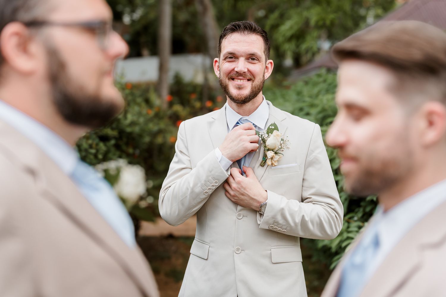 Groom in beige suit adjusting tie while smiling at groomsmen outdoors at wedding ceremony.