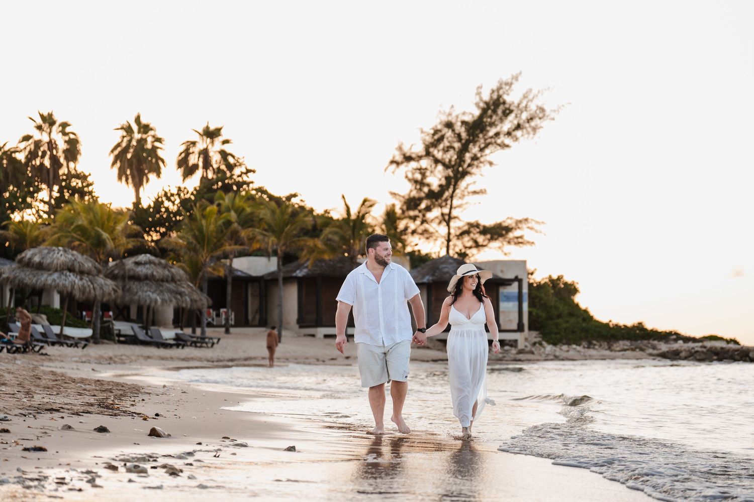 Two people walk hand in hand along a tropical beach at sunset with palm trees and thatched huts in the background.