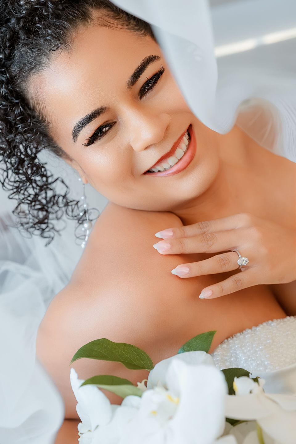 Smiling bride with curly hair wearing a diamond engagement ring, white veil, and holding white flowers.