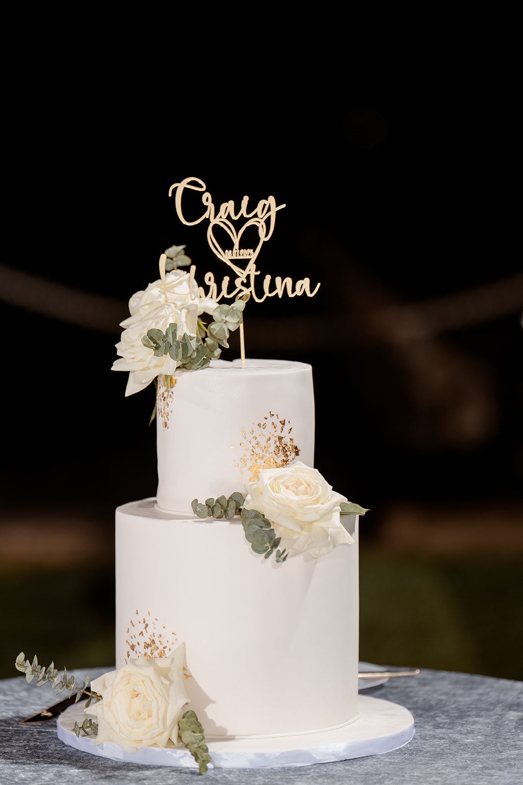 White two-tiered wedding cake with fresh roses and eucalyptus, topped with a gold script cake topper on a stone surface.