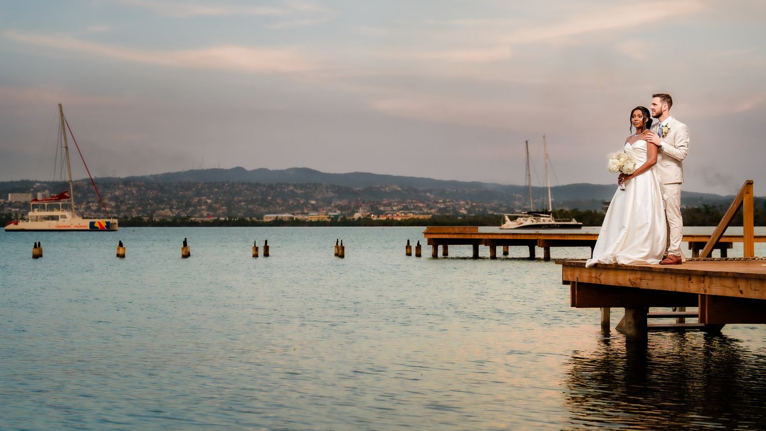 A bride and groom embrace on a wooden dock at sunset, with sailboats and a scenic waterfront town in the background.