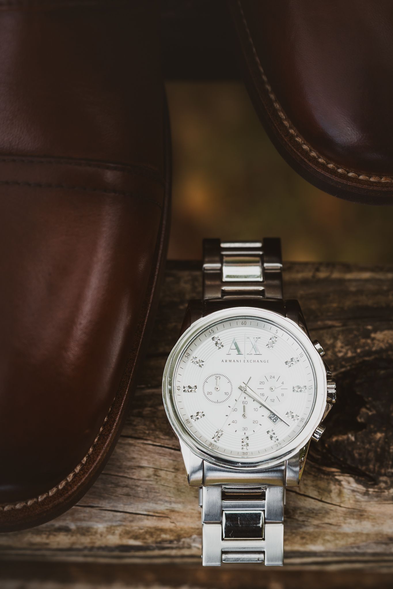 Silver Armani Exchange chronograph watch displayed between brown leather dress shoes on rustic wood surface.