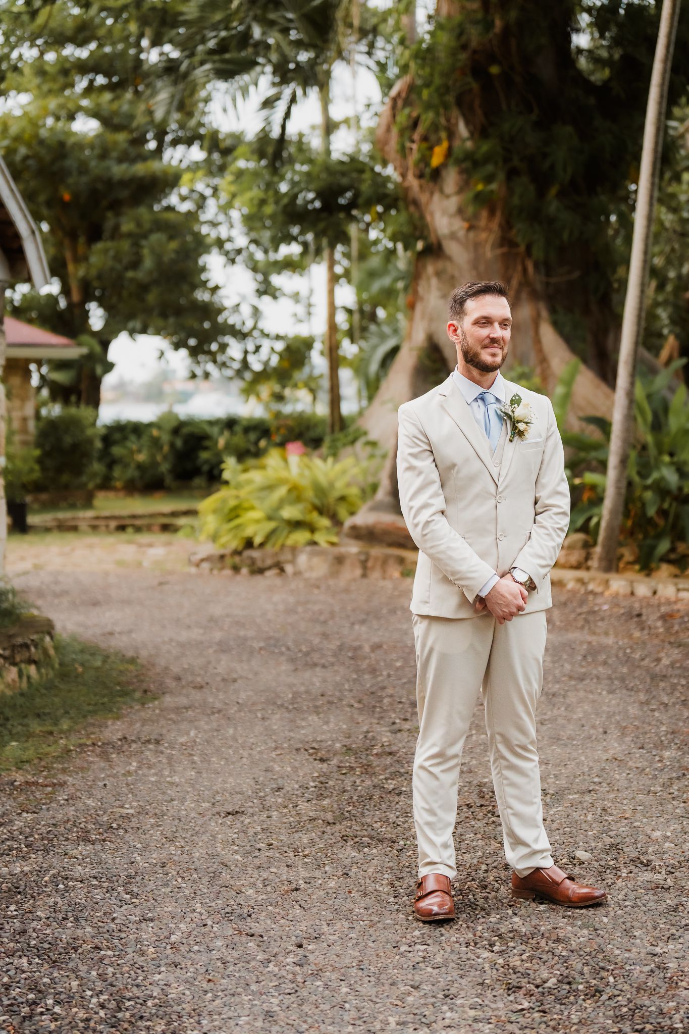 Groom in a beige suit with boutonniere smiling outdoors at a tropical wedding venue.