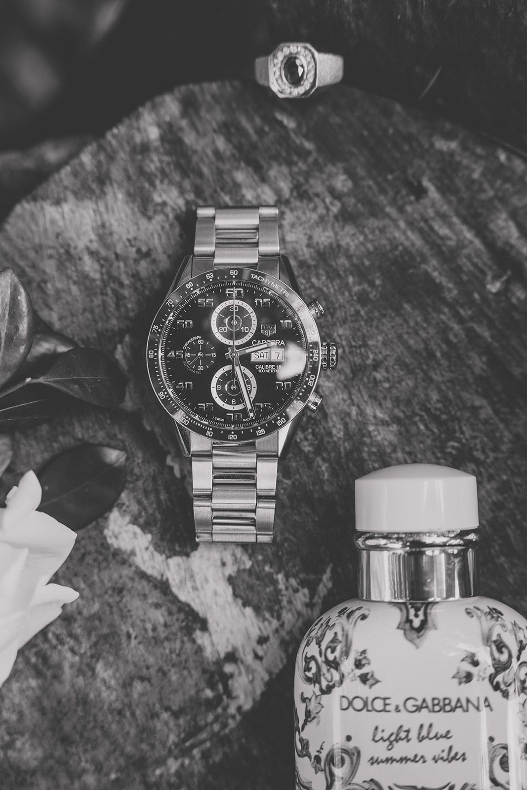 Black and white photo of a luxury watch, ring, and designer perfume bottle arranged on a textured surface.