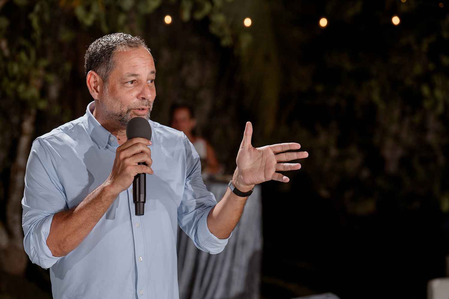 A person in a light colored shirt gestures while speaking into a microphone during a performance or presentation.