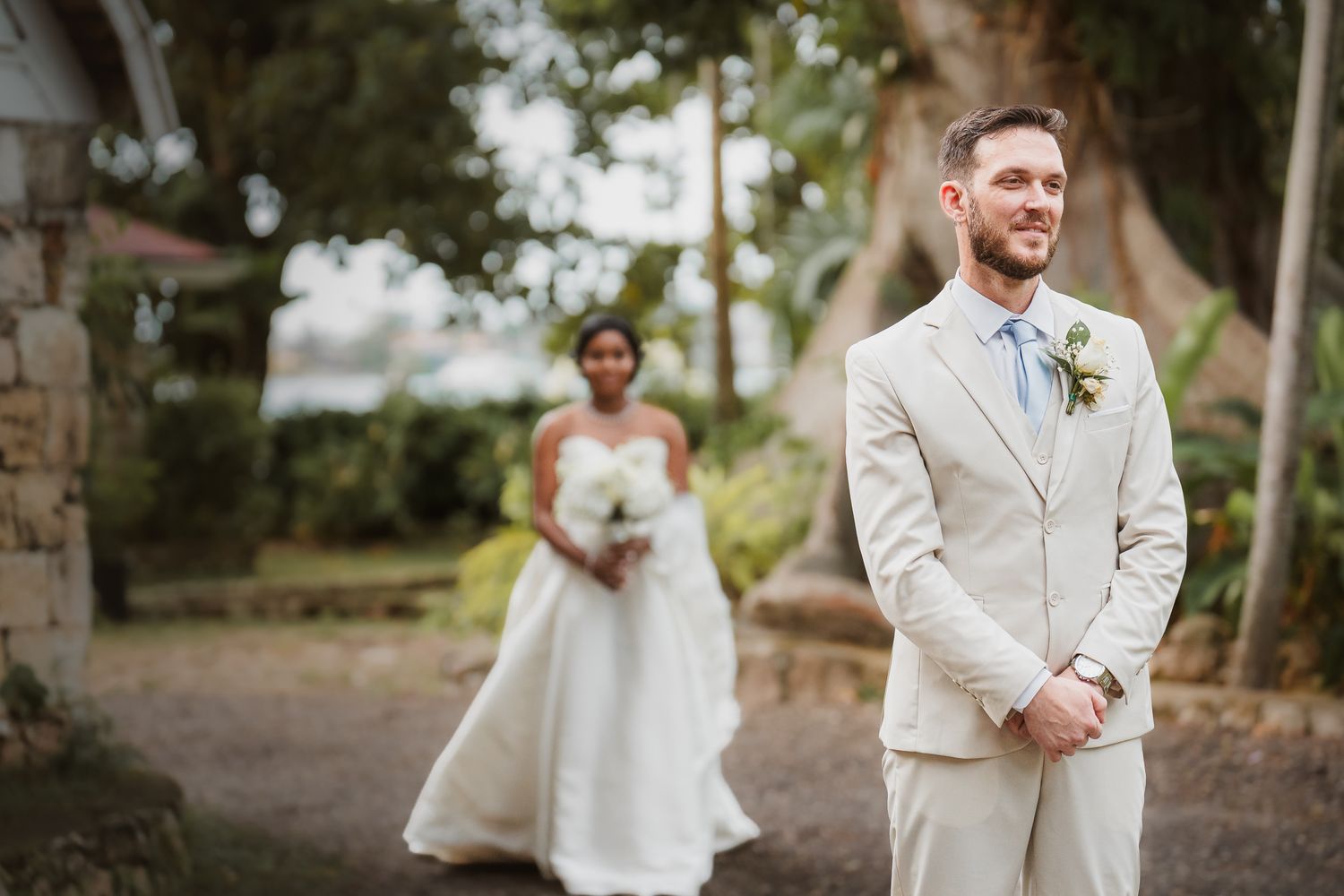 Groom in beige suit smiles during first look as bride in white gown approaches outdoors at wedding venue.