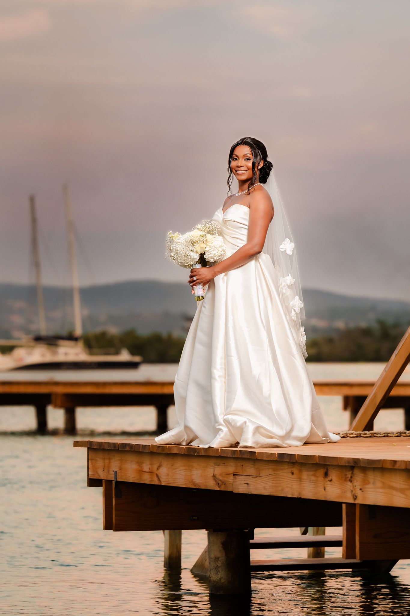 Bride in white gown holds bouquet on wooden dock at sunset with marina and hills in background.