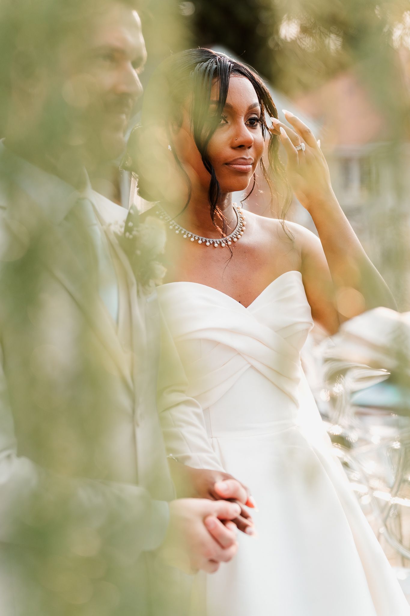 Bride in strapless white gown and diamond necklace holds groom's hand outdoors, framed by soft green foliage.