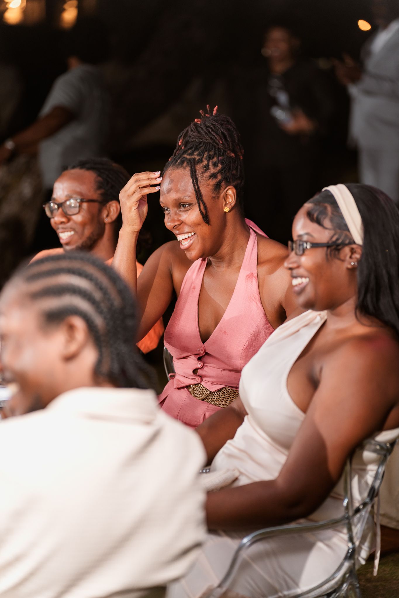 Smiling Black women in elegant dresses enjoying a celebration event, with one in pink halter dress laughing joyfully.