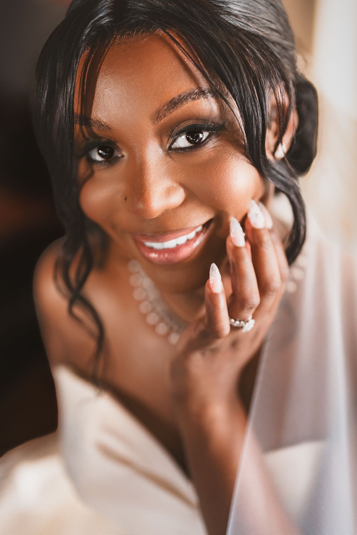 Smiling Black bride in white dress with pearl necklace and engagement ring, resting hand on cheek.