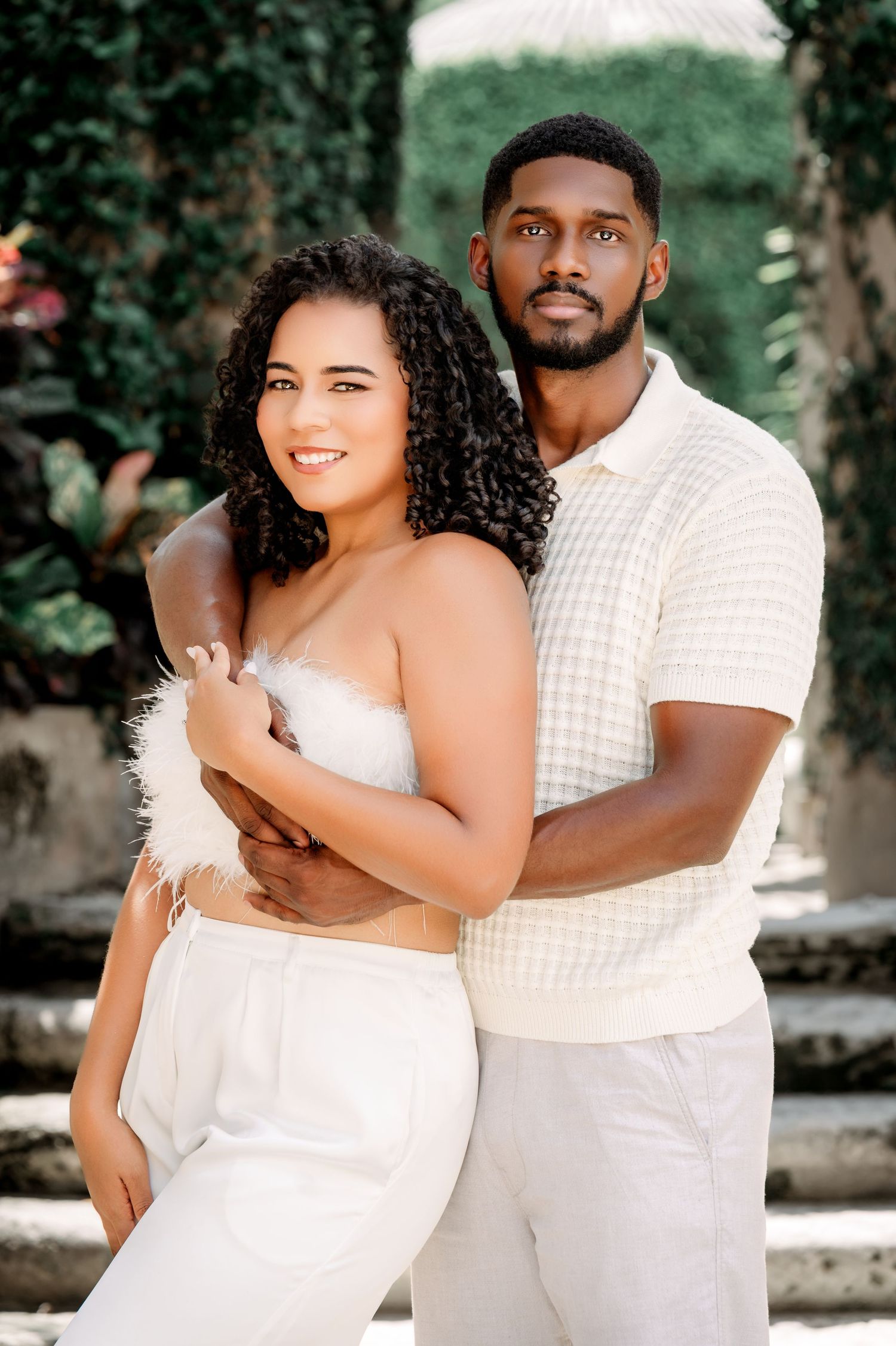 A couple in white outfits embraces outdoors in a romantic photoshoot with natural greenery in the background.