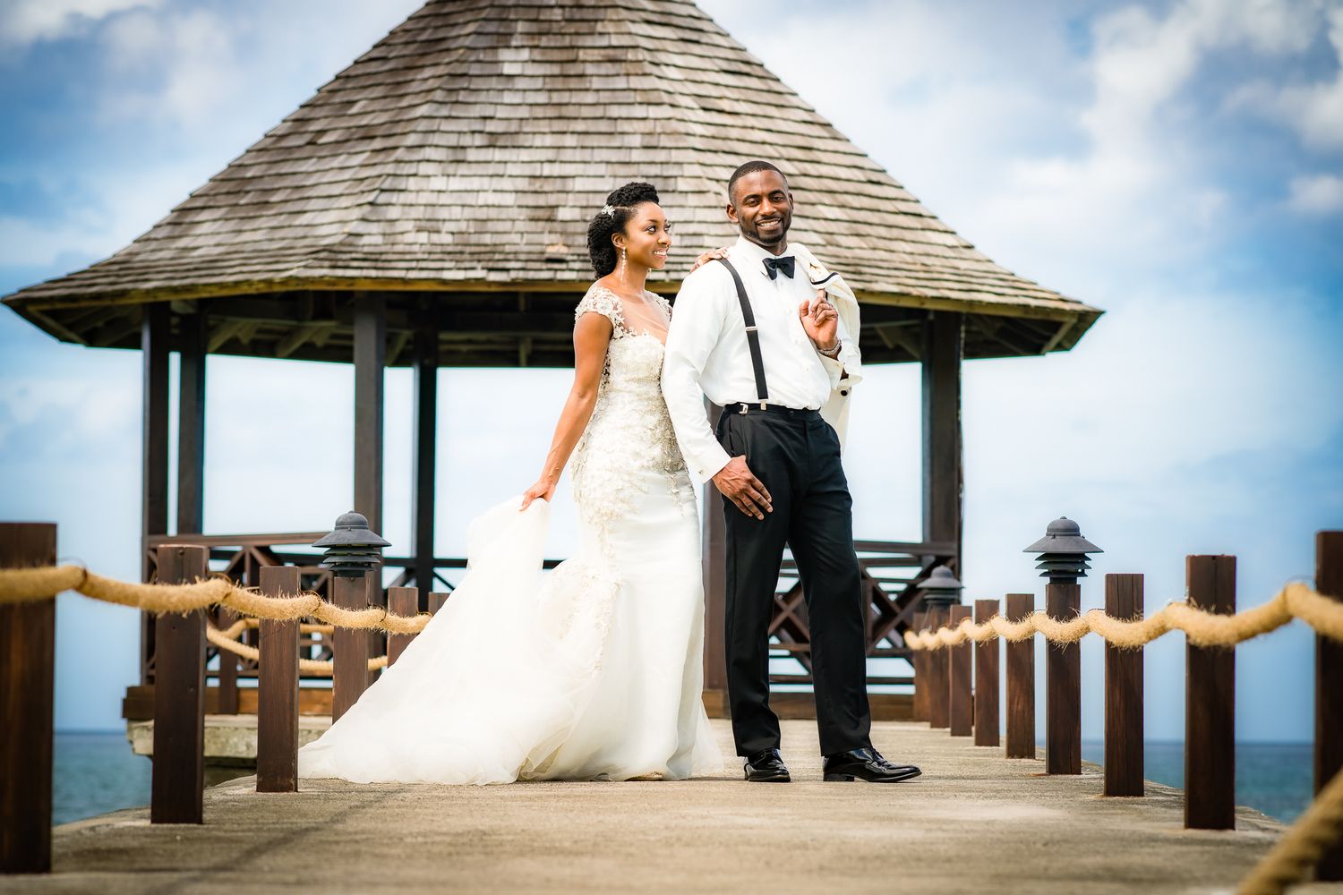 Bride and groom pose on a waterfront pier with a gazebo backdrop on their tropical wedding day.