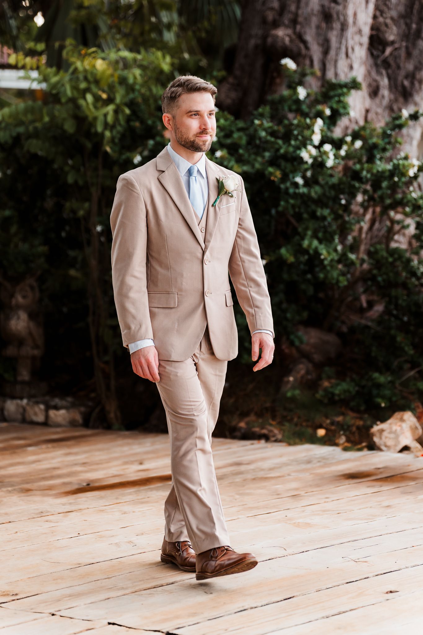 Groom in beige linen suit with blue tie and boutonniere walks on wooden deck outdoors.