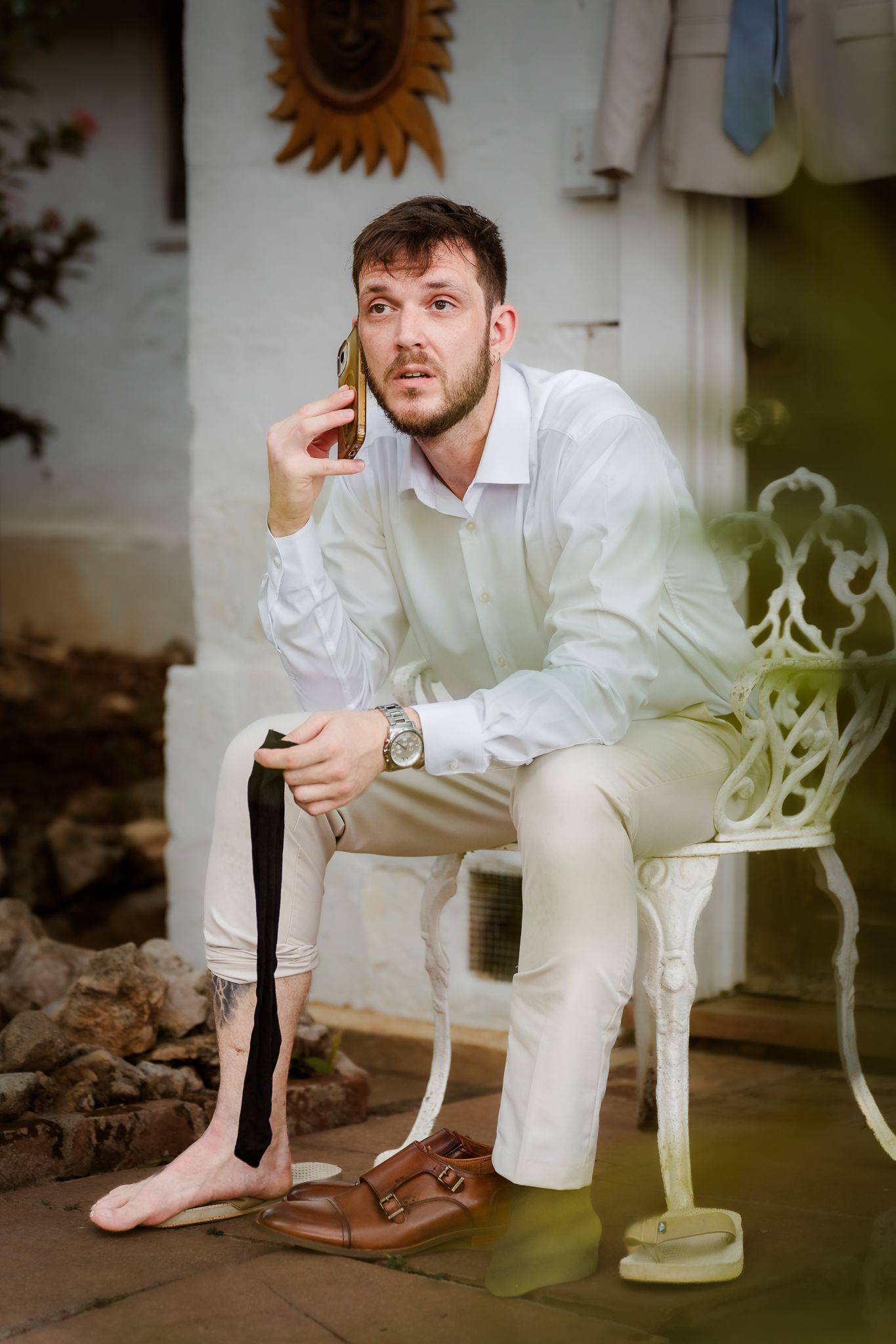 Man in white shirt and pants sitting on ornate chair, holding black tie and talking on phone outdoors.