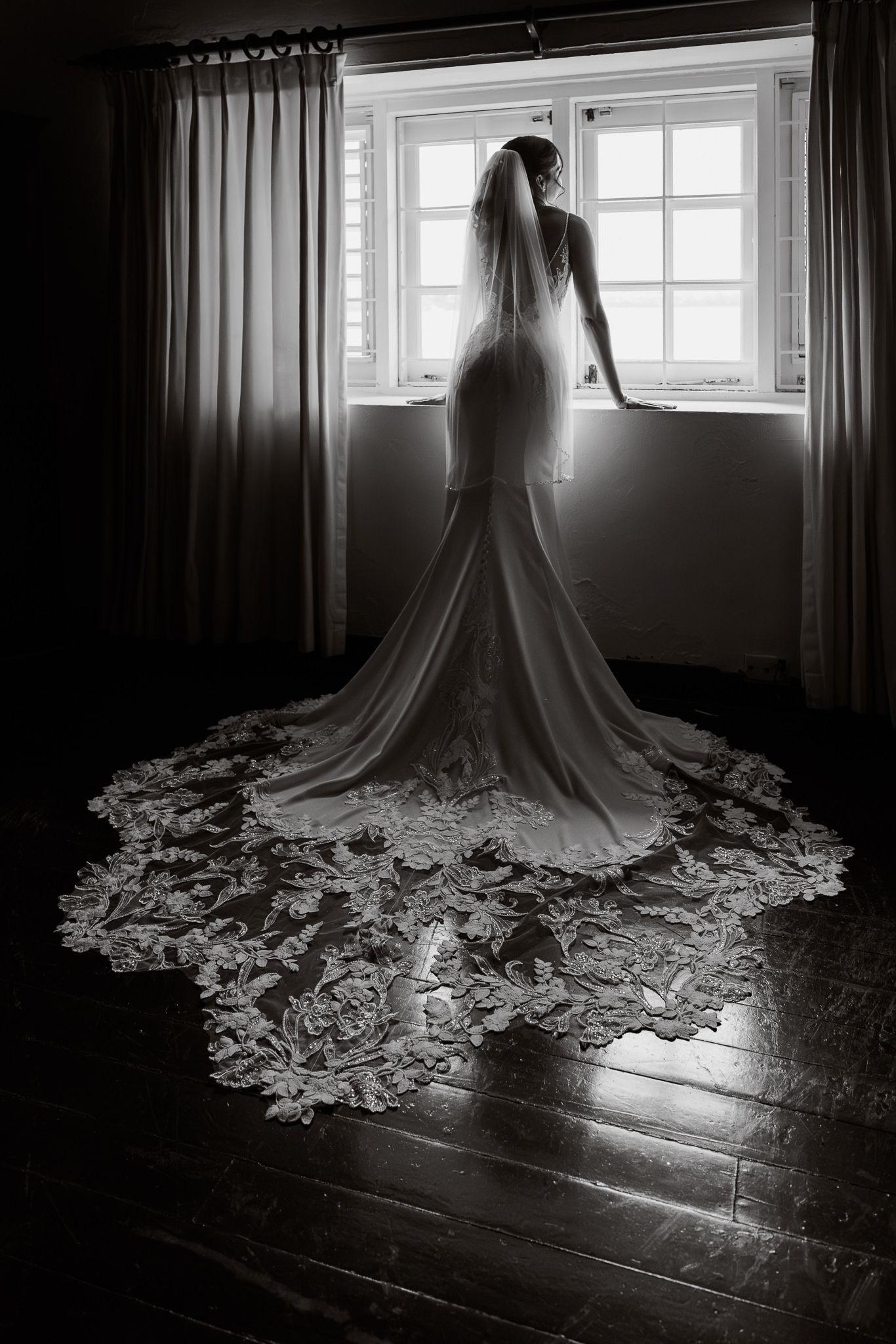 Silhouetted figure in a wedding dress stands by window creating dramatic contrast with flowing lace train on wooden floor.