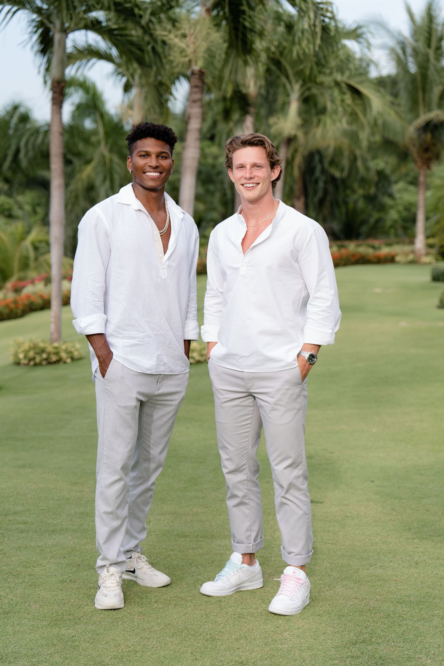 Two people wearing matching white shirts and light colored pants pose together on a golf course with palm trees in background.